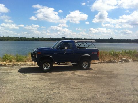 1982 Ford Bronco - A blue 1982 Ford Bronco is parked on a paved area next to a body of water under a partly cloudy sky.
