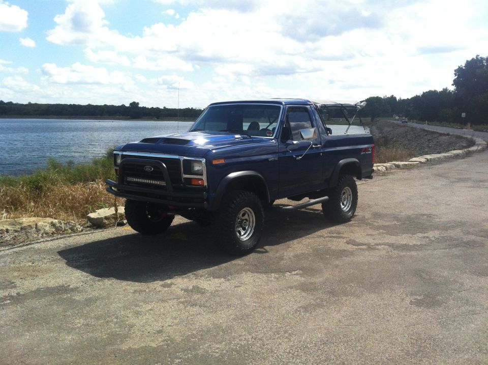 1982 Ford Bronco parked on asphalt near a body of water, with a blue sky and clouds