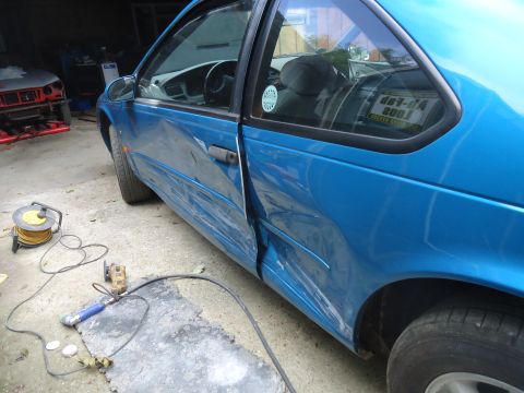 1994 Ford Thunderbird - This blue coupe shows significant damage to its driver's side door and fender, with repair tools visible nearby. Another vehicle is in the…