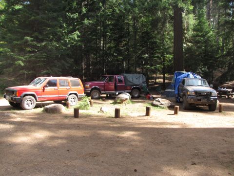1992 Ford F-150 - A campsite scene with multiple vehicles including a Jeep Cherokee, a maroon pickup truck with a tarp, and a dark-colored pickup truck.