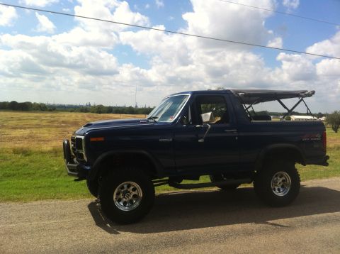 1982 Ford Bronco - A dark blue Ford Bronco is parked on a dirt road with a field and cloudy sky in the background.