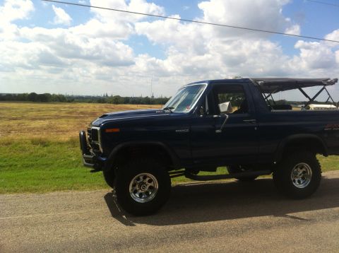 1982 Ford Bronco - A dark blue Ford Bronco is seen from the side, parked on a road with a field and distant city skyline in the background.