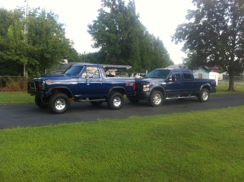 1982 Ford Bronco - Two blue Ford trucks, an older Bronco and a newer F-Series, are parked on a paved surface next to a grassy lawn.