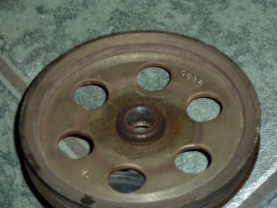 Rusty metal accessory pulley with six holes and central hub, resting on green tile floor