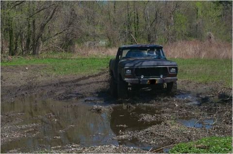 1979 Ford Bronco - A dark-colored Ford truck is navigating through a muddy, off-road terrain with trees in the background.