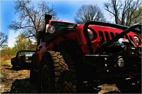 1979 Ford Bronco - Red Jeep Wrangler with snorkel and winch parked on a dirt road, another vehicle in background.