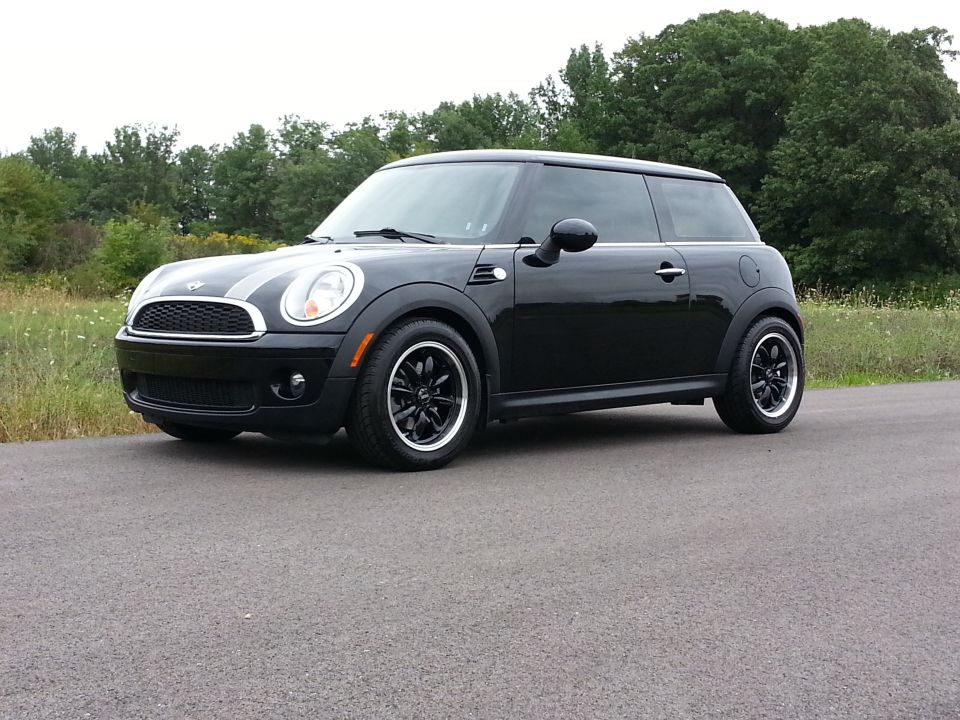 Black MINI Cooper parked on asphalt road, trees in background. Front 3/4 view