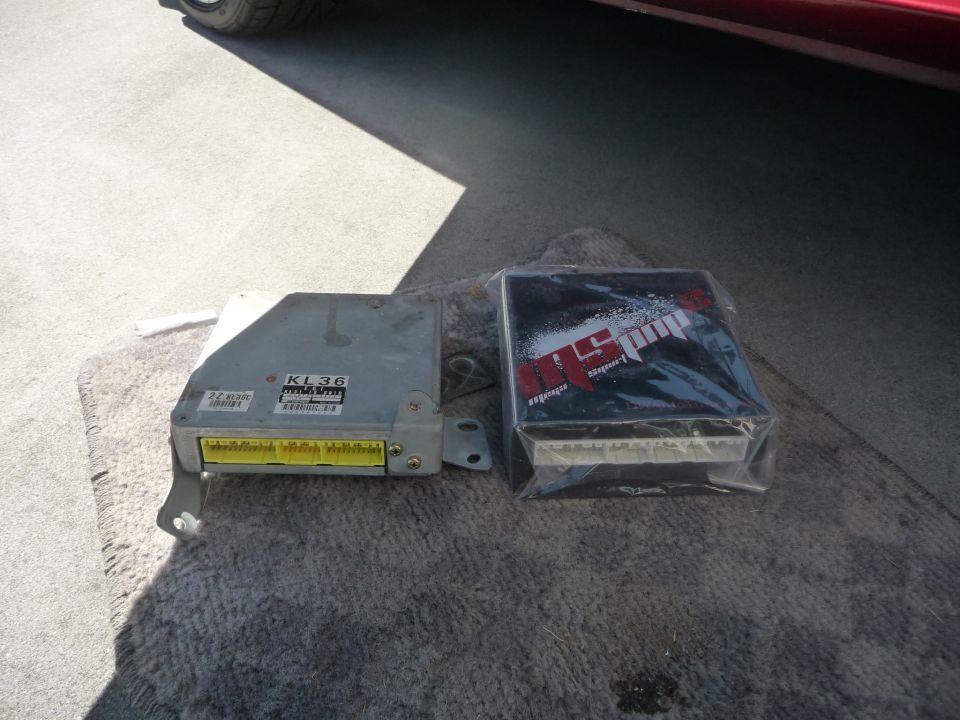 Two engine control units, one labeled KL36, sit on a rug next to a red car