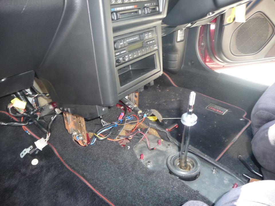 Car interior showing center console removed, exposed wiring, and shifter assembly. Floor mat has red trim