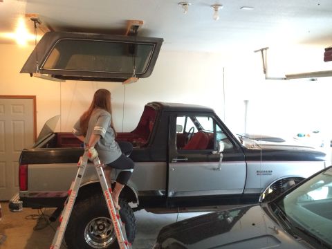 1989 Ford Bronco - A woman sits on a ladder next to a 1989 Ford Bronco, with its removable top stored on a hoist system above.