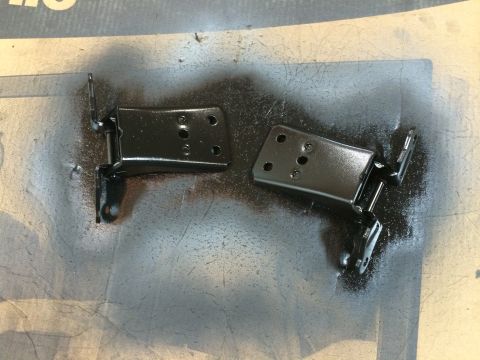 1989 Ford Bronco - Two black metal door hinges, recently painted, lie on a surface with overspray.