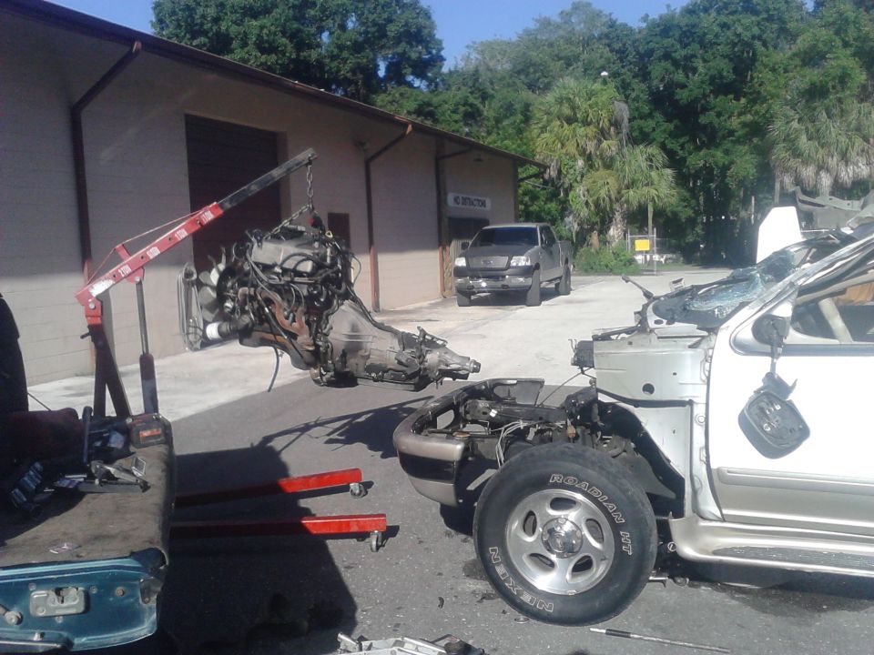 Engine and transmission on a red hoist being removed from a partially disassembled silver truck outside a garage