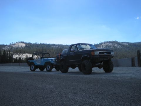 1989 Ford Bronco - A lifted black Ford Bronco and a blue Jeep CJ are parked side-by-side on a gravel lot with a mountainous forest in the background.