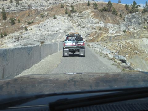 1989 Ford Bronco - A light blue and white Ford Bronco with a roof rack loaded with gear drives on a mountain road.