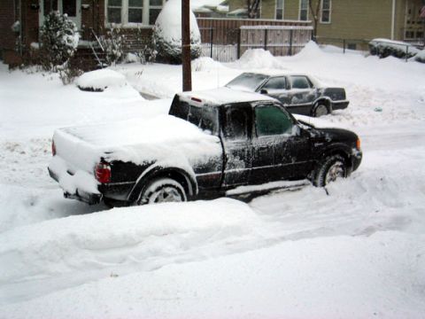 1996 Ford Crown Victoria - Two vehicles are partially buried in snow on a street, with a house and fence in the background.