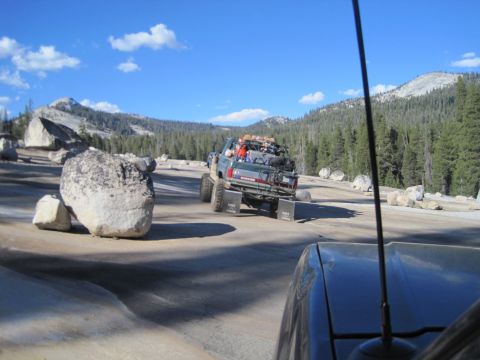 1989 Ford Bronco - Multiple off-road vehicles travel in convoy on a dirt road surrounded by rocks and pine trees under a blue sky.