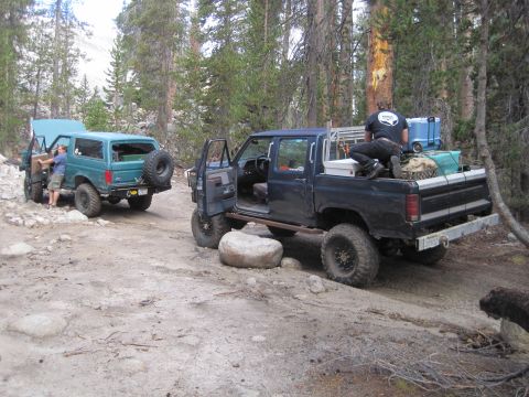 1989 Ford Bronco - Two off-road vehicles are stopped on a dirt path surrounded by trees.