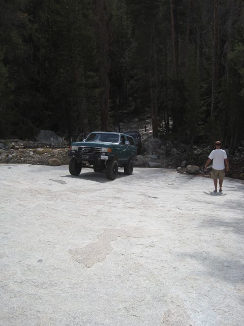 1989 Ford Bronco - A green Ford Bronco is parked in a wooded area, with another vehicle behind it and a person standing nearby.