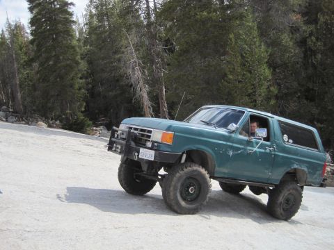 1989 Ford Bronco - A green Ford Bronco drives on a dirt road surrounded by trees.