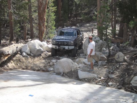 1989 Ford Bronco - A dark pickup truck navigates a challenging rocky path in a wooded area, with text on the windshield indicating 'DUSTY 9-2014'.