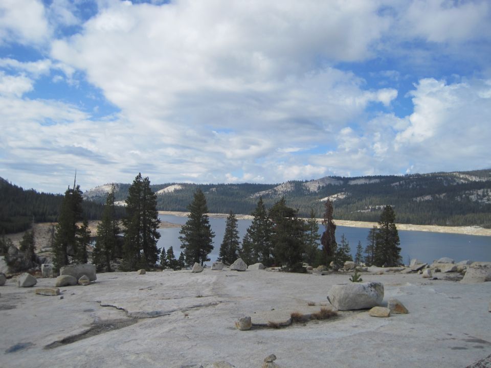 View of a calm lake surrounded by pine trees and mountains under a cloudy sky