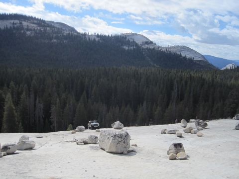 1989 Ford Bronco - A lone off-road vehicle is parked on a rocky plateau, surrounded by a vast forest and distant mountains under a cloudy sky.