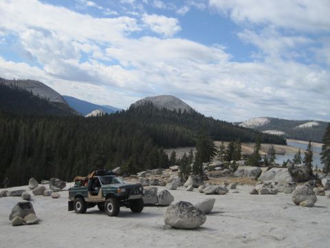 1989 Ford Bronco - A green 1989 Ford Bronco is parked on a rocky surface with a forest and lake in the background under a cloudy sky.
