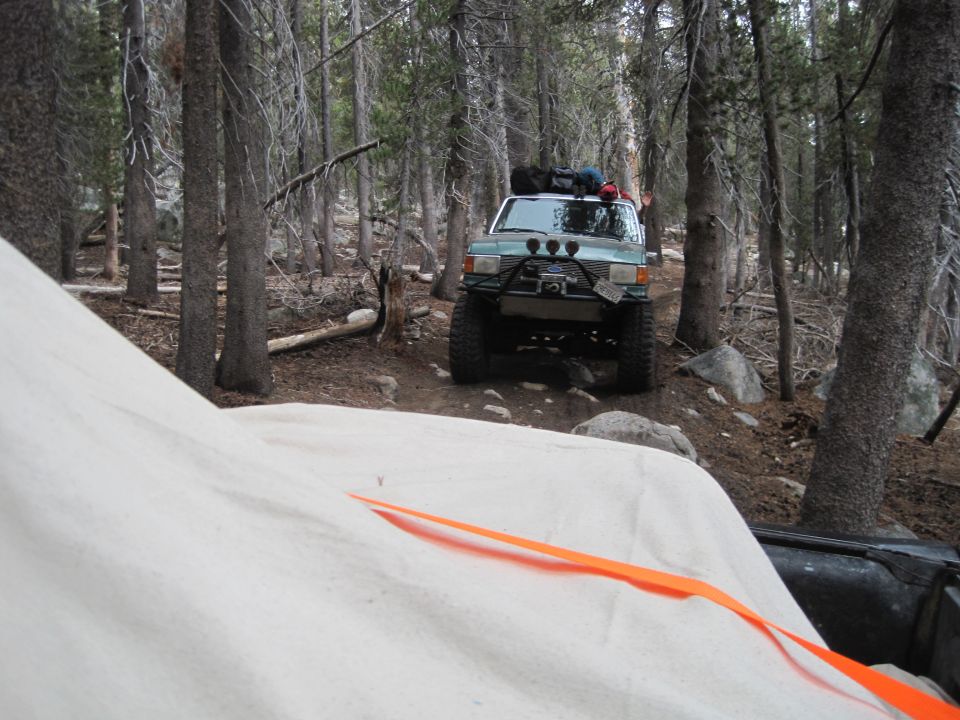 Light blue SUV with off-road tires, winch, and roof rack parked on a dirt path in a forest