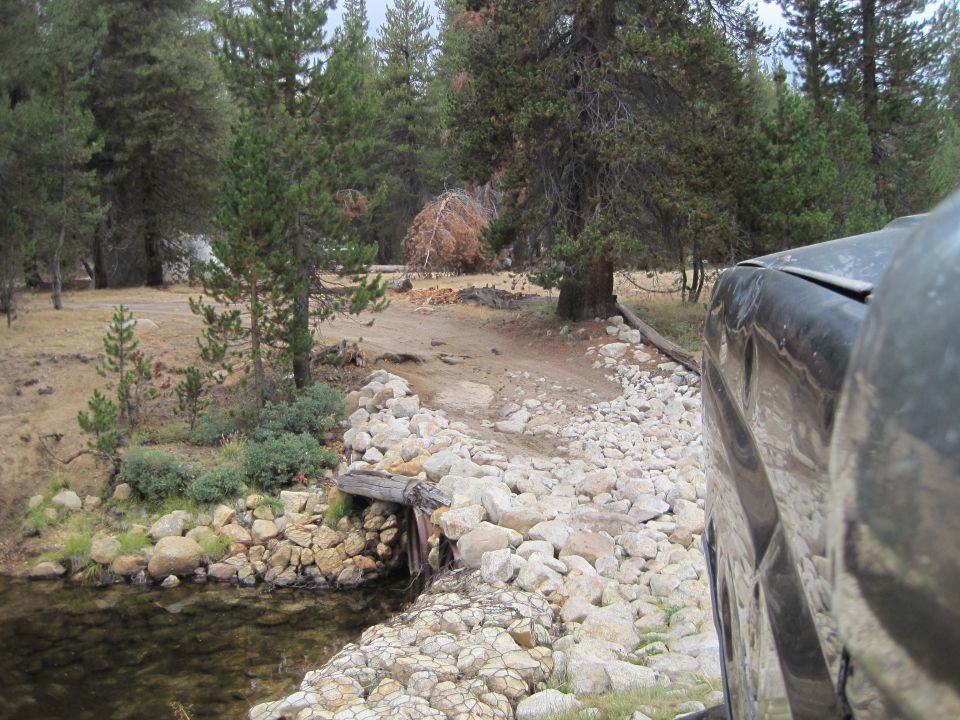 View from inside a vehicle driving on a rocky path next to a stream. Trees and brush surround the area