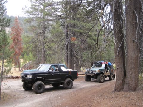 1989 Ford Bronco - Two off-road vehicles drive down a dirt path in a forest, one black Bronco and one lighter colored SUV.