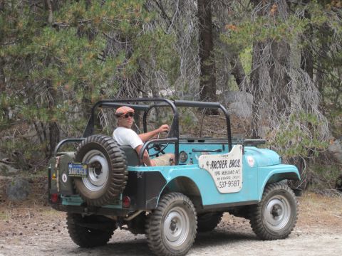 1989 Ford Bronco - A man drives a blue Jeep off-road, with a spare tire mounted on the rear and company signage on the door.