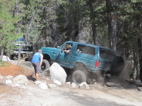 1989 Ford Bronco - A teal Ford Bronco is shown off-roading on a dirt path with rocks and trees surrounding it.