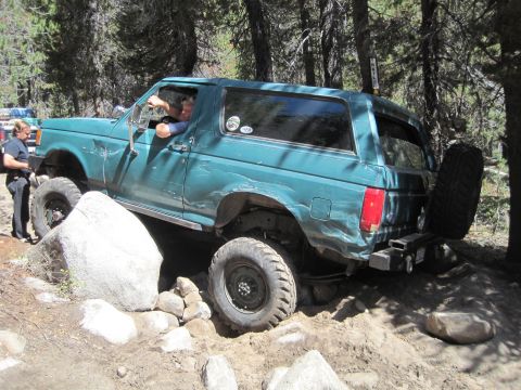 1989 Ford Bronco - A teal SUV is shown off-roading on a dirt trail surrounded by trees, with a person driving.