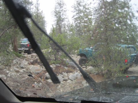 1989 Ford Bronco - Two SUVs traverse a challenging, rocky path through a dense forest, with raindrops on the windshield.