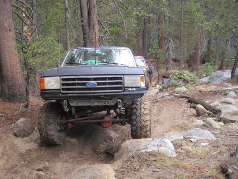 1989 Ford Bronco - A black 1989 Ford Bronco navigates a challenging off-road trail with large rocks and trees surrounding it.
