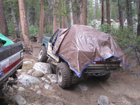 1989 Ford Bronco - Two off-road vehicles are parked in a wooded area with one covered by a tarp.