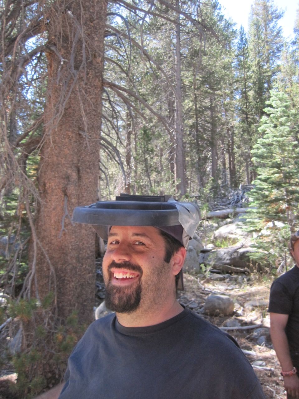 Man in a dark t-shirt smiles, wearing a black trash can lid on his head, in a forest setting