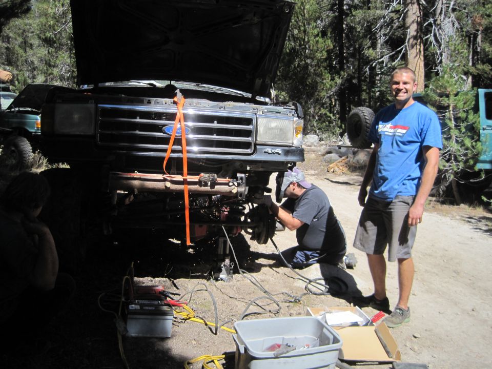 Ford truck with hood up, people working on front suspension. Another green vehicle visible