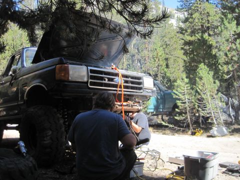 1989 Ford Bronco - Mechanics work on a lifted black Ford F-Series truck in a wooded area, with another green truck visible in the background.