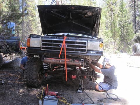 1989 Ford Bronco - A person works on the front end of a black Ford truck with its hood up, while another vehicle is partially visible to the left.