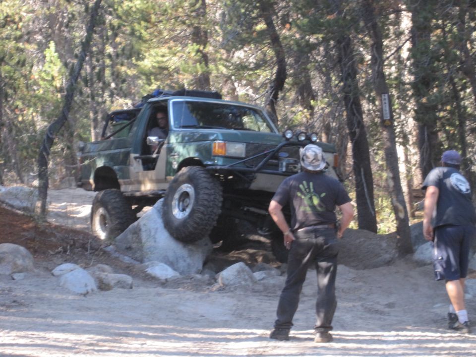 Green off-road vehicle with large tires climbing over rocks on a dirt trail. Two men stand nearby