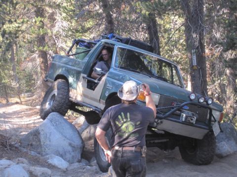 1989 Ford Bronco - A man in a hat offers a beer to the driver of a green Ford Bronco navigating a rocky trail.