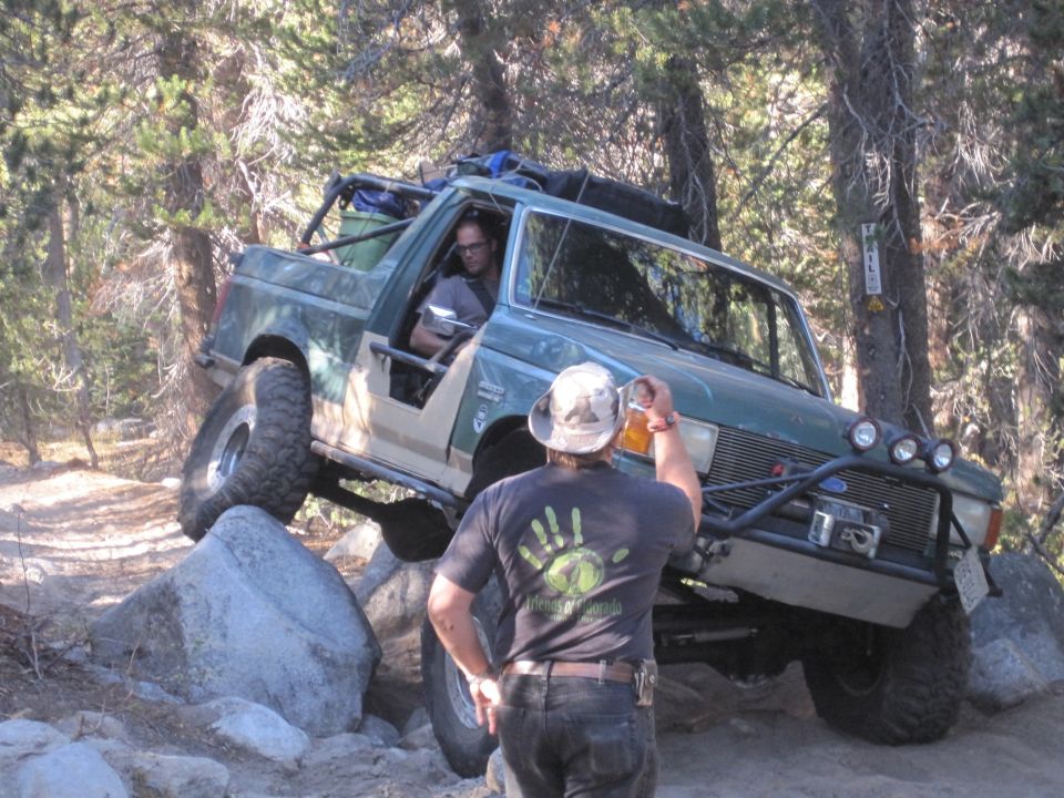 Green Ford Bronco off-roading over rocks, driver holding beer