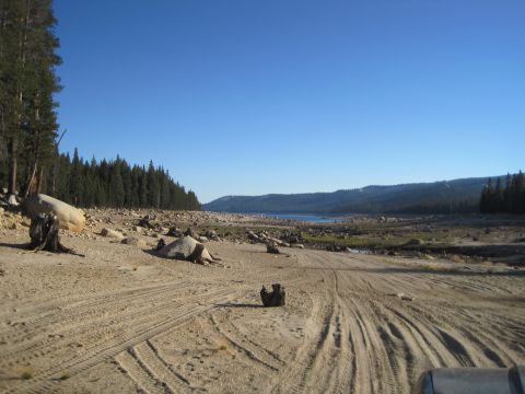 1989 Ford Bronco - A dry, rocky landscape with tire tracks leading towards a distant lake, bordered by a dense forest.
