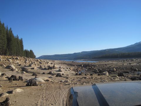 1989 Ford Bronco - A dry lakebed with numerous rocks and tree stumps stretches towards a distant body of water under a clear blue sky.
