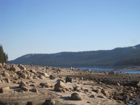 1989 Ford Bronco - A lone off-road vehicle navigates a , arid landscape near a body of water under a clear blue sky.