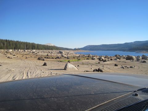 1989 Ford Bronco - The view from inside a vehicle shows a dry lake bed dotted with rocks and tree stumps, leading to a distant blue lake and forested hills.