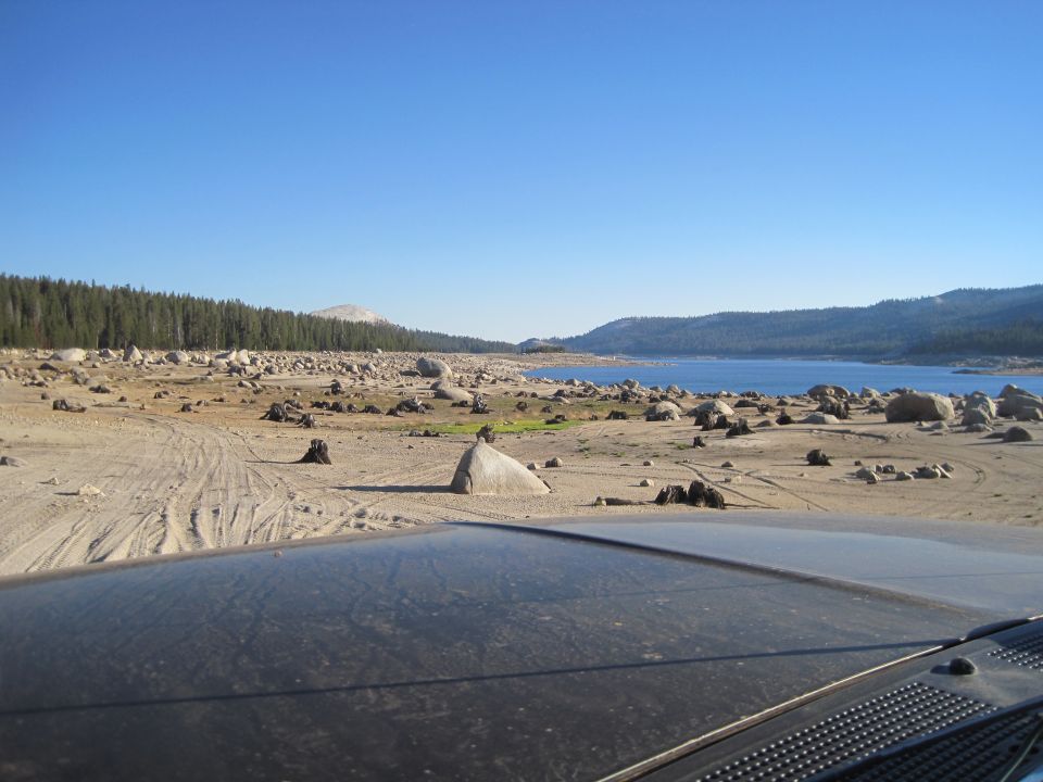 View from car interior of a dry lake bed with rocks and trees under a clear blue sky