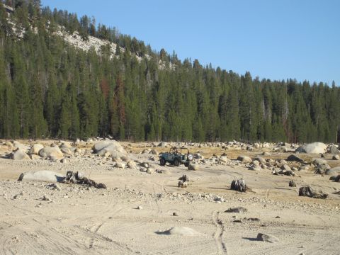 1989 Ford Bronco - An off-road vehicle sits in a barren landscape with a dense forest and rocky hillside behind it.