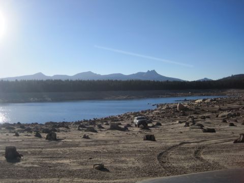 1989 Ford Bronco - An SUV sits on a barren, rocky shore next to a body of water, with a forested mountain range under a clear blue sky.